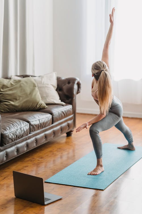 Free Full body side view of unrecognizable female in activewear practicing extended side angle posture while performing yoga exercise in living room with laptop Stock Photo