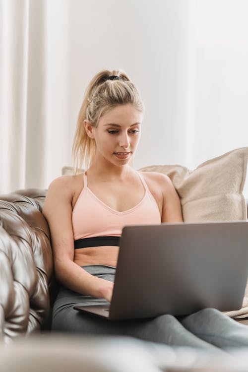 Concentrated young lady in sportive bra and leggings browsing on netbook and sitting on couch with pillow in light living room