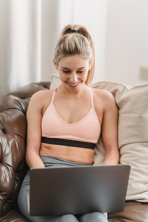 Happy young lady in sportive bra and leggings browsing on netbook while sitting on leather couch with pillow in light room