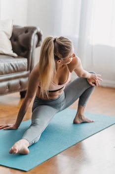 Full body young female in sportive bra and leggings doing exercises on blue yoga mat near leather sofa near curtains in bright living room and looking away