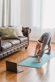 A woman in a yoga pose at home, exercising on a mat with a laptop nearby.