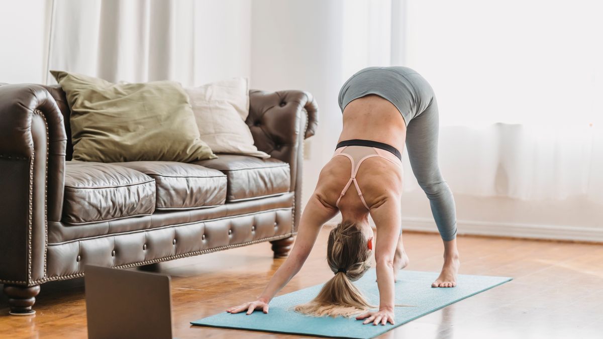 Full length of faceless lady wearing leggings and sportive bra stretching back on yoga mat near opened netbook and leather couch with pillows in light living room near curtains