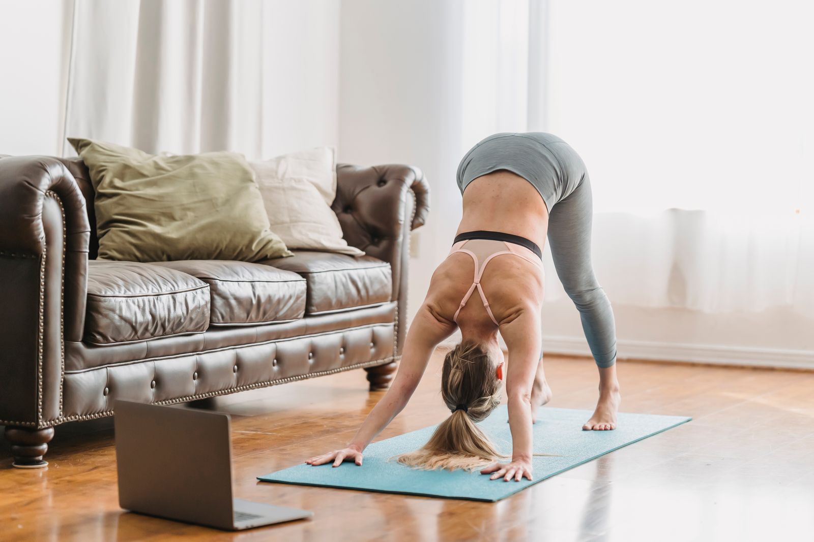 Woman practicing downward-facing dog pose on a yoga mat at home near a laptop and couch