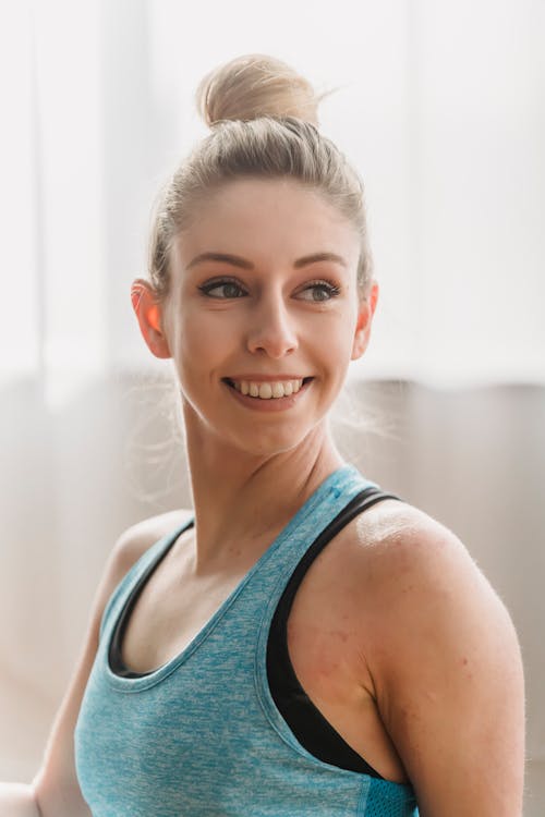 Cheerful woman in activewear smiling and looking away while sitting in light room at home while preparing for yoga training