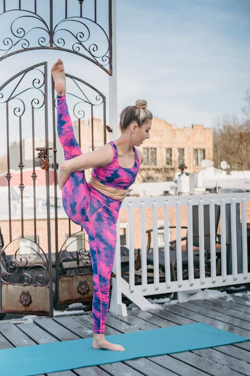 Full body of slim female in sportswear standing on one leg while practicing yoga on mat against buildings in city