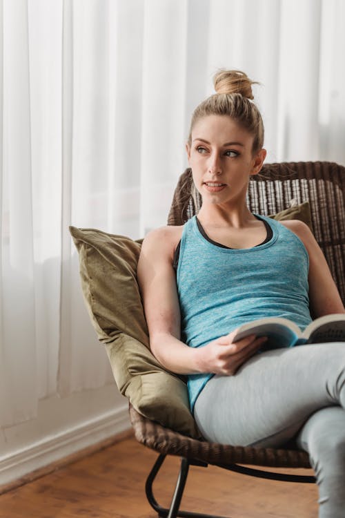 Relaxed female sitting on pillow in comfortable braided armchair with book in hands and looking away after training