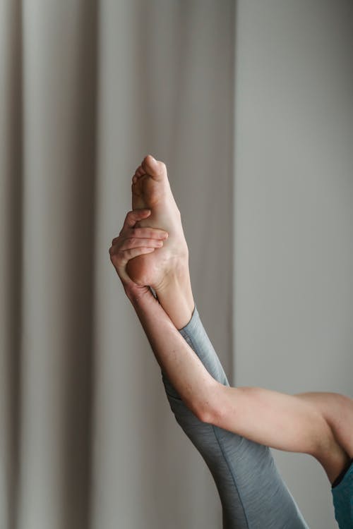 Crop unrecognizable barefoot female practicing stretching exercise by raising straight leg up with hand and pointing toes in room with gray curtains