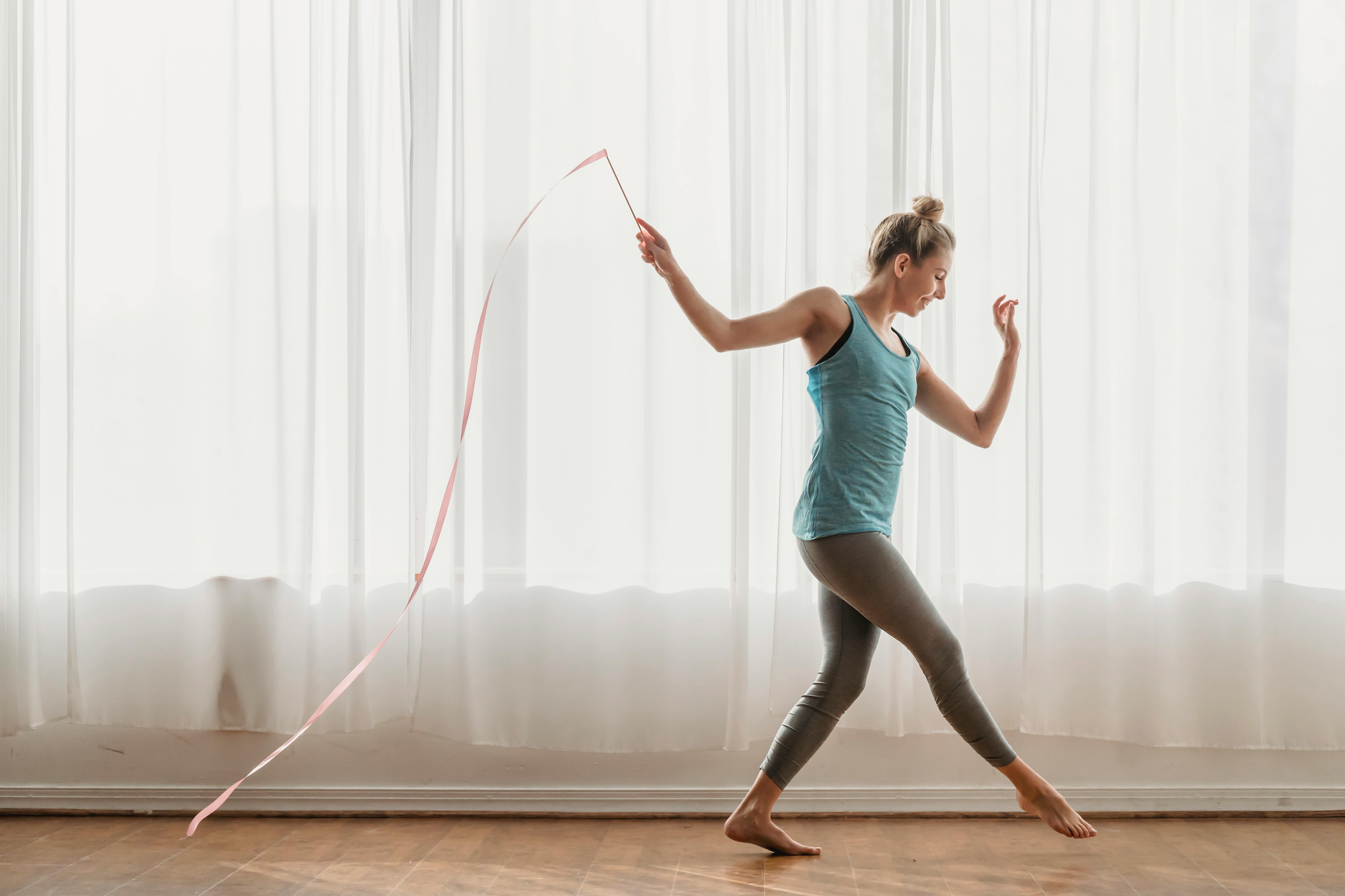 Free Side view of slender young female athlete doing steps with pink ribbon in hand behind back along full wall window Stock Photo