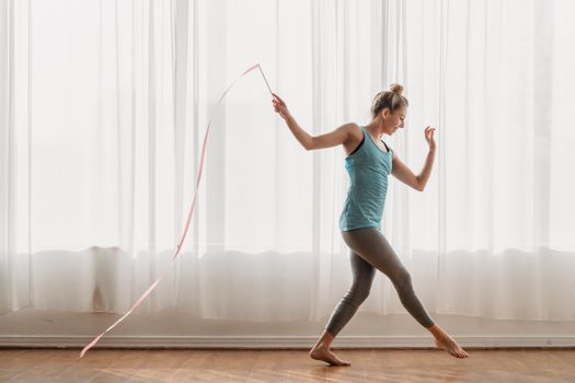 Side view of slender young female athlete doing steps with pink ribbon in hand behind back along full wall window