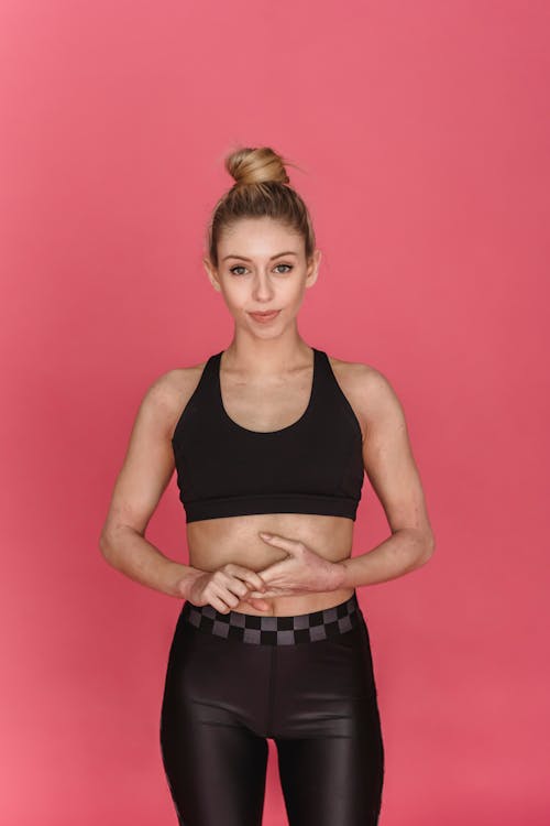 Young female athlete wearing black sports bra and leggings standing in studio against pink background and looking at camera