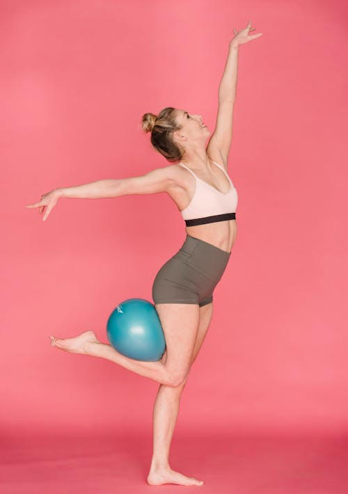 Side view full body of graceful positive female athlete outstretching arms while standing with fitness ball on leg against pink background
