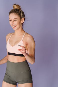 Positive slim muscular female wearing sportive top and shorts standing on purple background while standing in light room of studio
