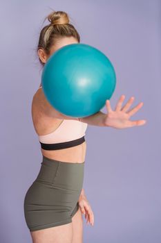 Faceless fit female in sportive top and shorts practicing with fitness ball while standing on purple background in modern studio