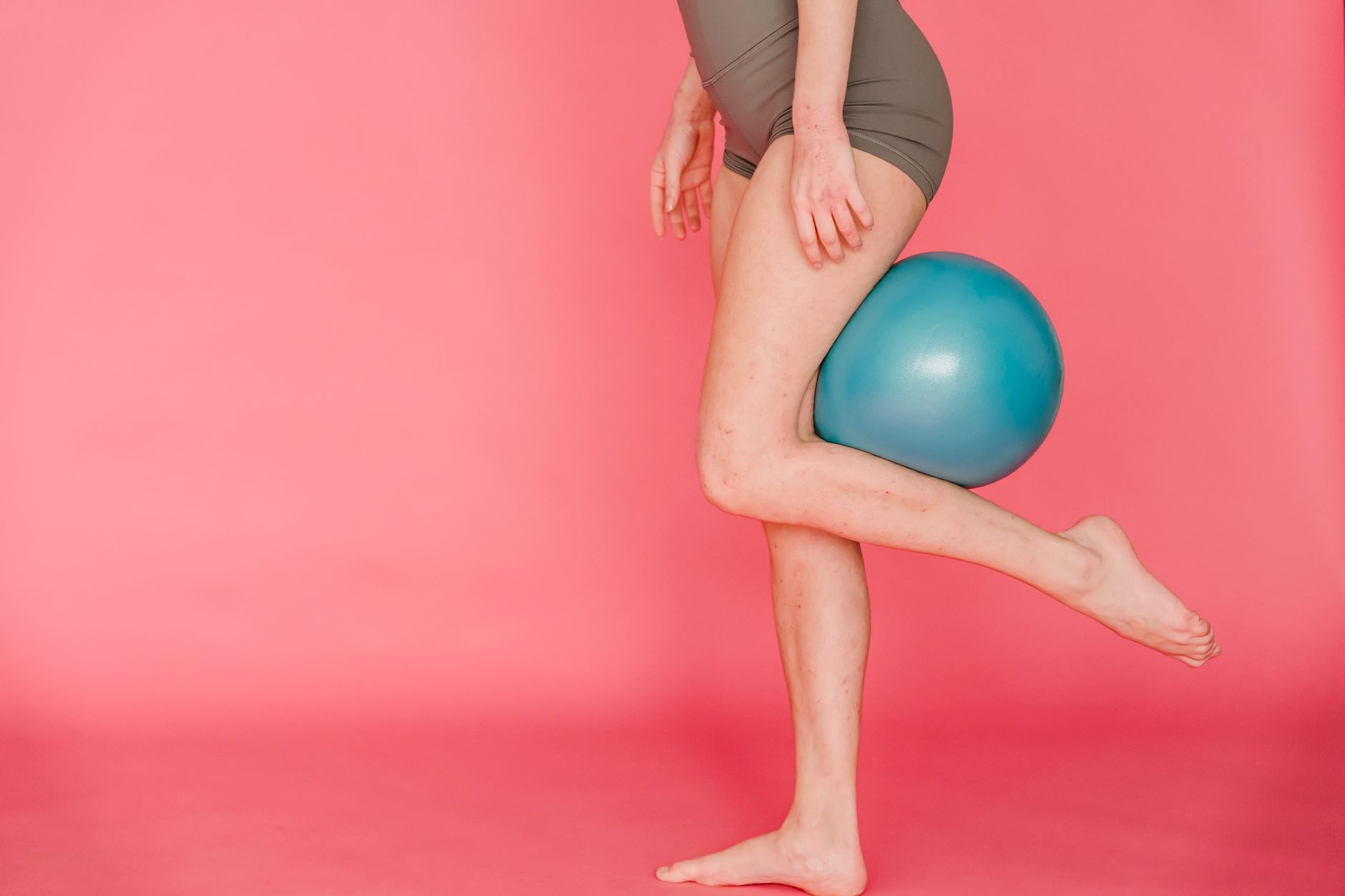 Side view of unrecognizable barefoot slim female gymnast wearing sportive shorts doing exercise with fitness ball while standing on pink background in studio