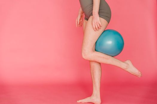 Side view of unrecognizable barefoot slim female gymnast wearing sportive shorts doing exercise with fitness ball while standing on pink background in studio
