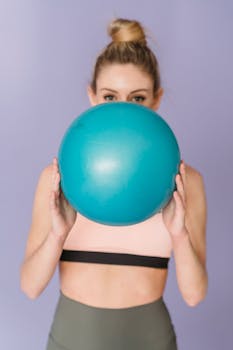 Sportive female wearing activewear looking at camera while covering half of face with fit ball on purple background in studio