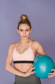 Confident woman holding a fitness ball in a purple-hued studio setting.
