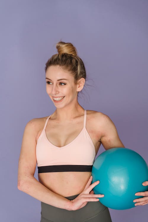 Cheerful muscular female in sportswear smiling and looking away while standing on purple background with fitness ball during training
