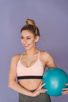 Cheerful muscular female in sportswear smiling and looking away while standing on purple background with fitness ball during training