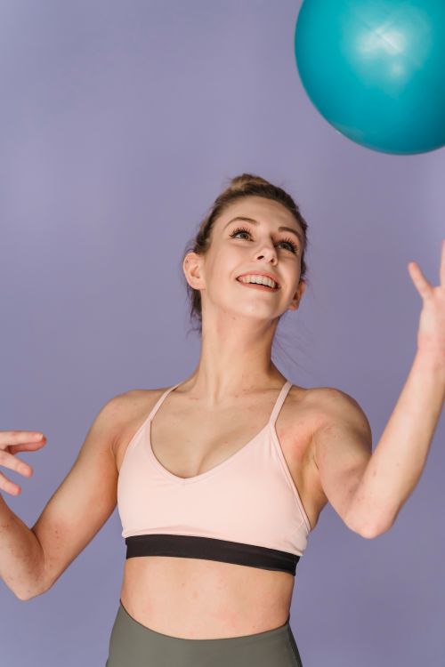 Free Cheerful sportive female in activewear looking up and throwing fitness ball high in air on purple background during workout in studio Stock Photo