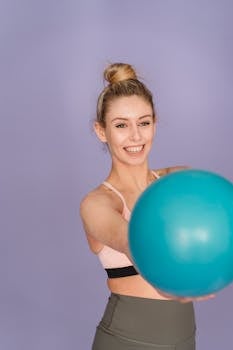 Cheerful slender female in sportswear showing blue shiny gym ball and smiling on purple background
