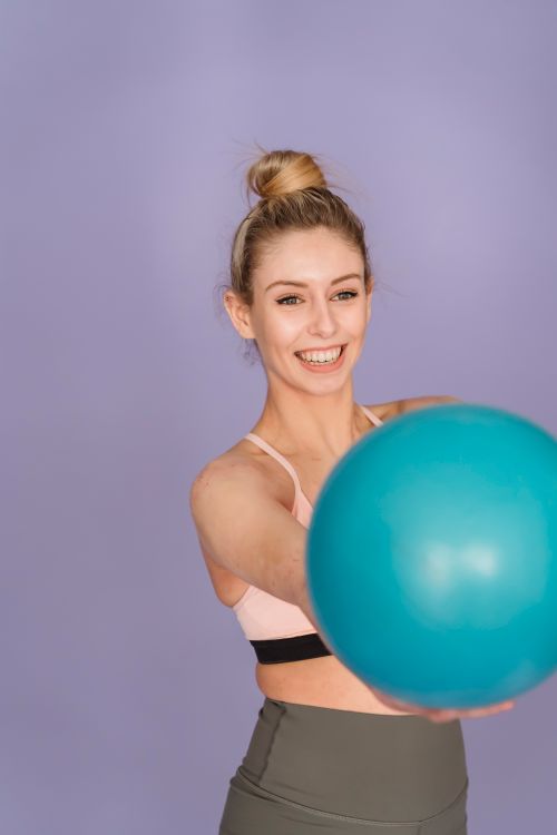 Free Cheerful slender female in sportswear showing blue shiny gym ball and smiling on purple background Stock Photo
