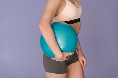Crop anonymous slim female in sportswear with blue gym ball on purple background