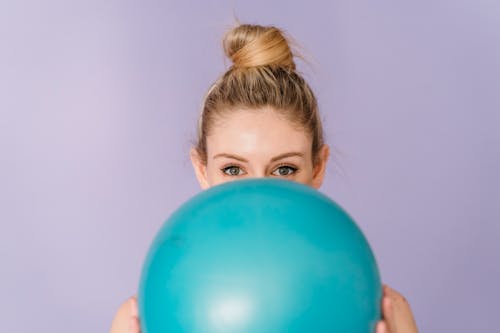 Cheerful anonymous female demonstrating blue shiny gym ball in hands on purple background of studio