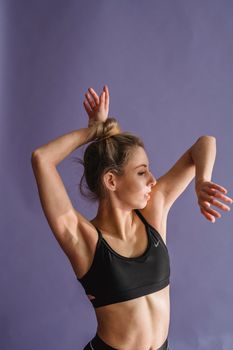 Pensive slim female in black sportswear standing with raised arms and dancing in studio against purple background