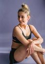 Full body of widely smiling slim female in black sportswear sitting on floor with bent knees and crossed arms looking away on sunny day in studio