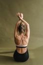 Back view of unrecognizable female in sportswear sitting in front of olive wall while doing yoga exercise