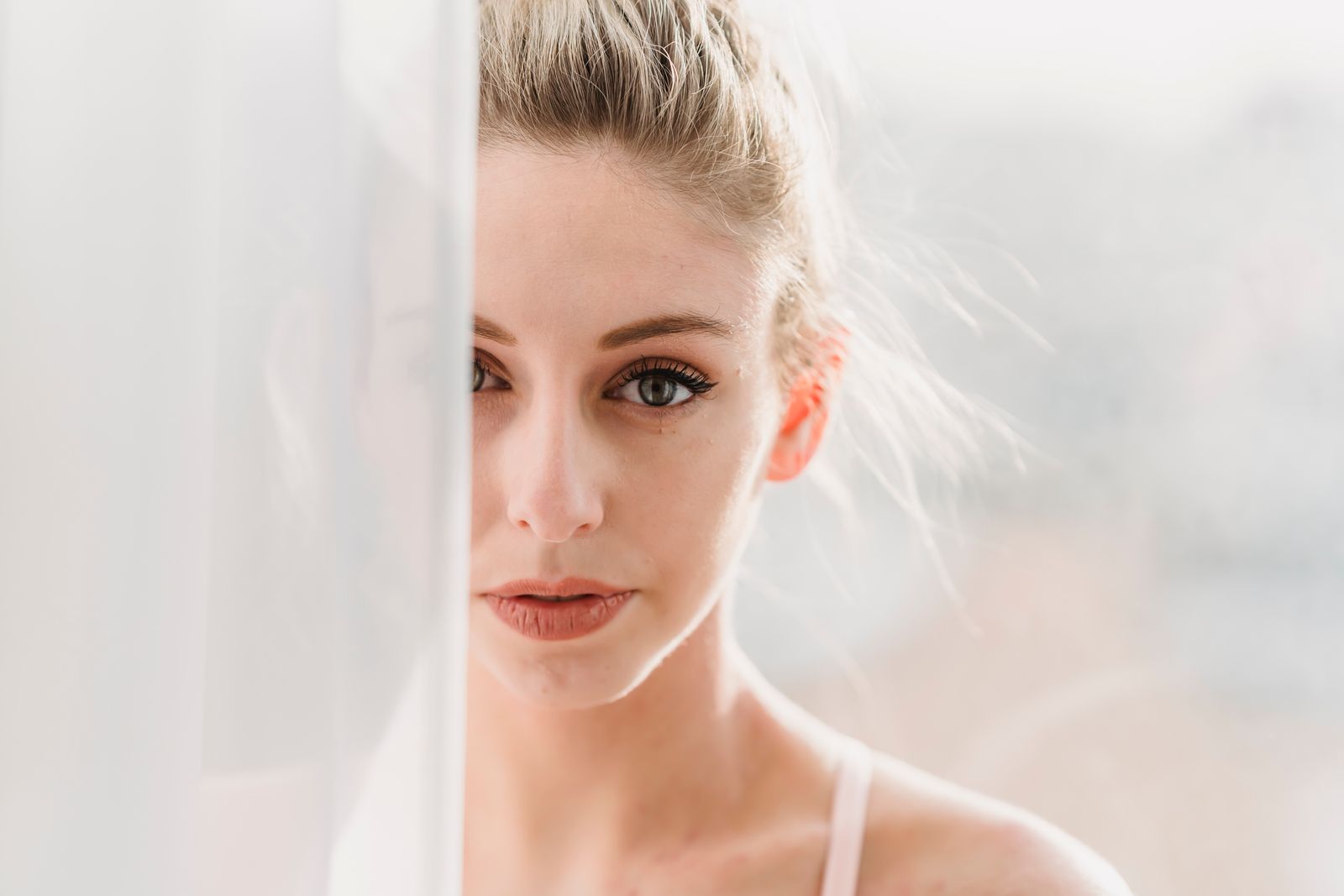 Woman with blond hair partly behind a sheer white curtain, looking at the camera in soft sunlight