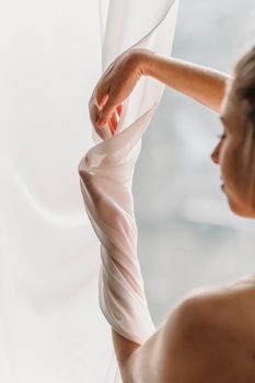 Side view of crop graceful young lady with bare shoulders touching white chiffon curtains in sunlight