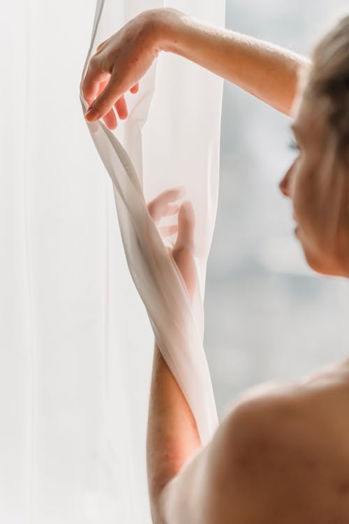 Side view of crop young feminine woman with bare shoulders touching white chiffon curtains gracefully near window in sunlight