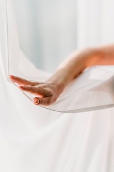 Crop anonymous lady touching soft white fabric near window on sunny day in light room