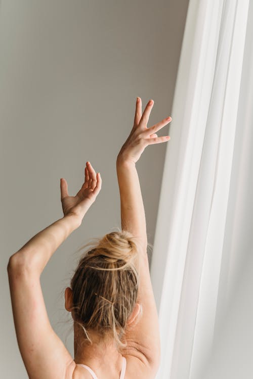 Free Back view of unrecognizable young lady with blond hair performing dance with hands above head near white curtains in daylight Stock Photo