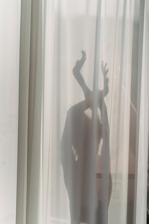 Shadow of anonymous female dancer performing sensual movements behind translucent white curtains in sunlight