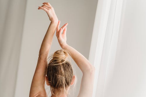 Back view of unrecognizable female with hair in bun standing with hands above head near white curtain in light room