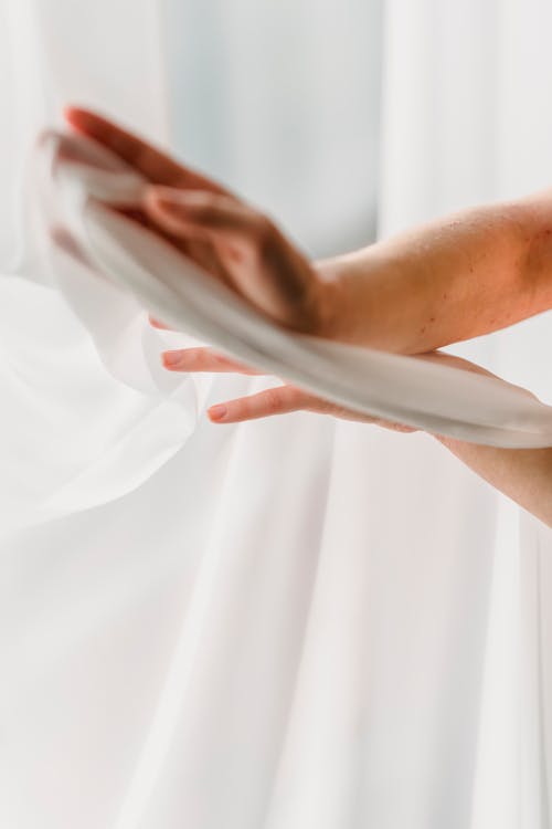 Hands of anonymous person touching gently white long elegant cloth while standing near window in modern light room at home