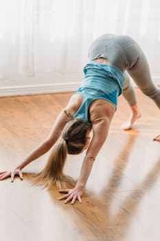 An adult woman performs the downward dog yoga pose on a wooden floor at home, highlighting fitness and wellness.
