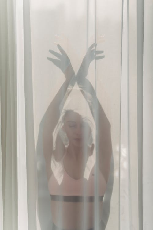 Calm crop female with closed eyes wearing top standing with hands above head behind light curtain in room with sunlight