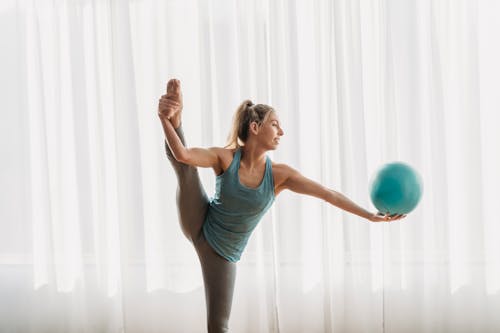 Smiling female athlete doing vertical split by raising straight leg with hand and looking at gymnastic ball on outstretched arm