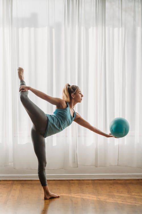 Side view of young slim woman in sportswear standing in front of window and training with gymnastic ball