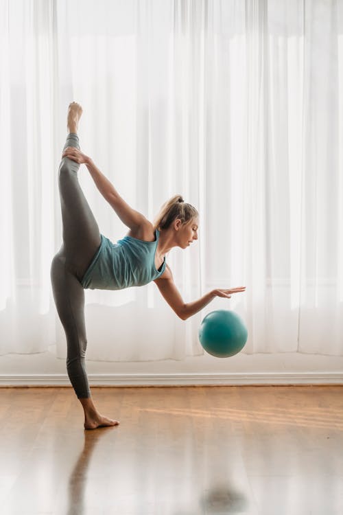 Free Side view of young female gymnast hitting rubber ball on parquet in light studio and balancing on leg Stock Photo