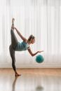 Side view of young female gymnast hitting rubber ball on parquet in light studio and balancing on leg