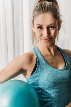 Cheerful female athlete in activewear standing in fitness studio with blue rubber ball and looking at camera