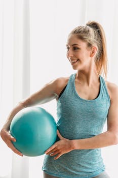 Positive slim female athlete in sportswear smiling and looking away while holding gymnastic ball in modern fitness studio