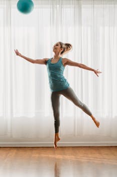 Smiling slim female in sportswear jumping up with fit ball spreading arms and stretching forefeet on sunny day in light room in dance studio
