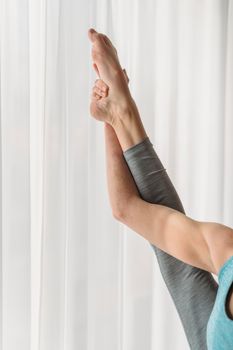 Anonymous female in sportswear stretching raised leg standing against white curtain in light room