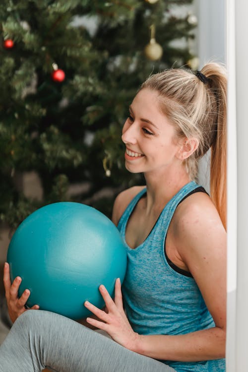 Free Smiling young female in sportswear holding fit ball in hands and looking away while sitting on floor on background of decorated Christmas tree Stock Photo